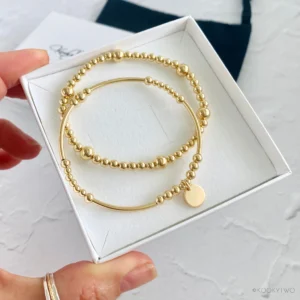 A hand holding a white box with two simple gold bracelets inside—one made of small round beads and the other with beads, a smooth bar, and a small round charm.